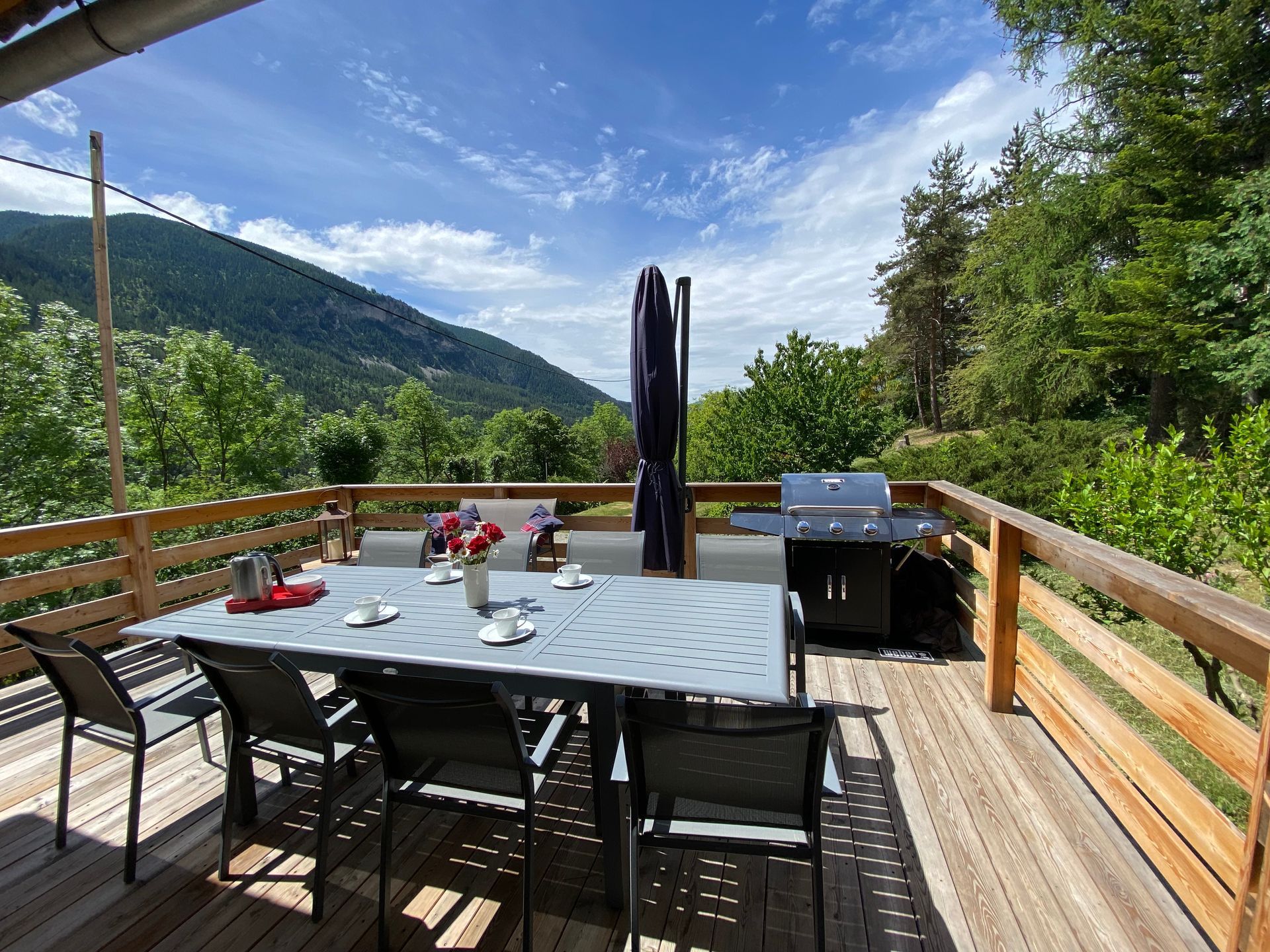 Terrasse en bois avec table à manger, chaises, barbecue et vue sur la montagne par une journée ensoleillée