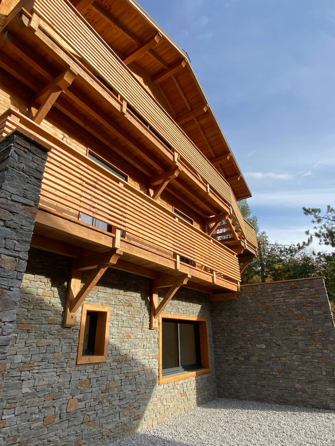 Un chalet en bois de deux étages, avec une partie en bois avec des balcons et un rez-de-chaussée en pierre.