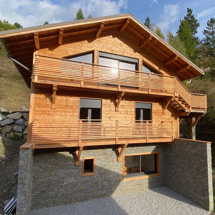 Un chalet en bois de deux étages avec balcons, construit sur des fondations en béton avec parement en pierre.