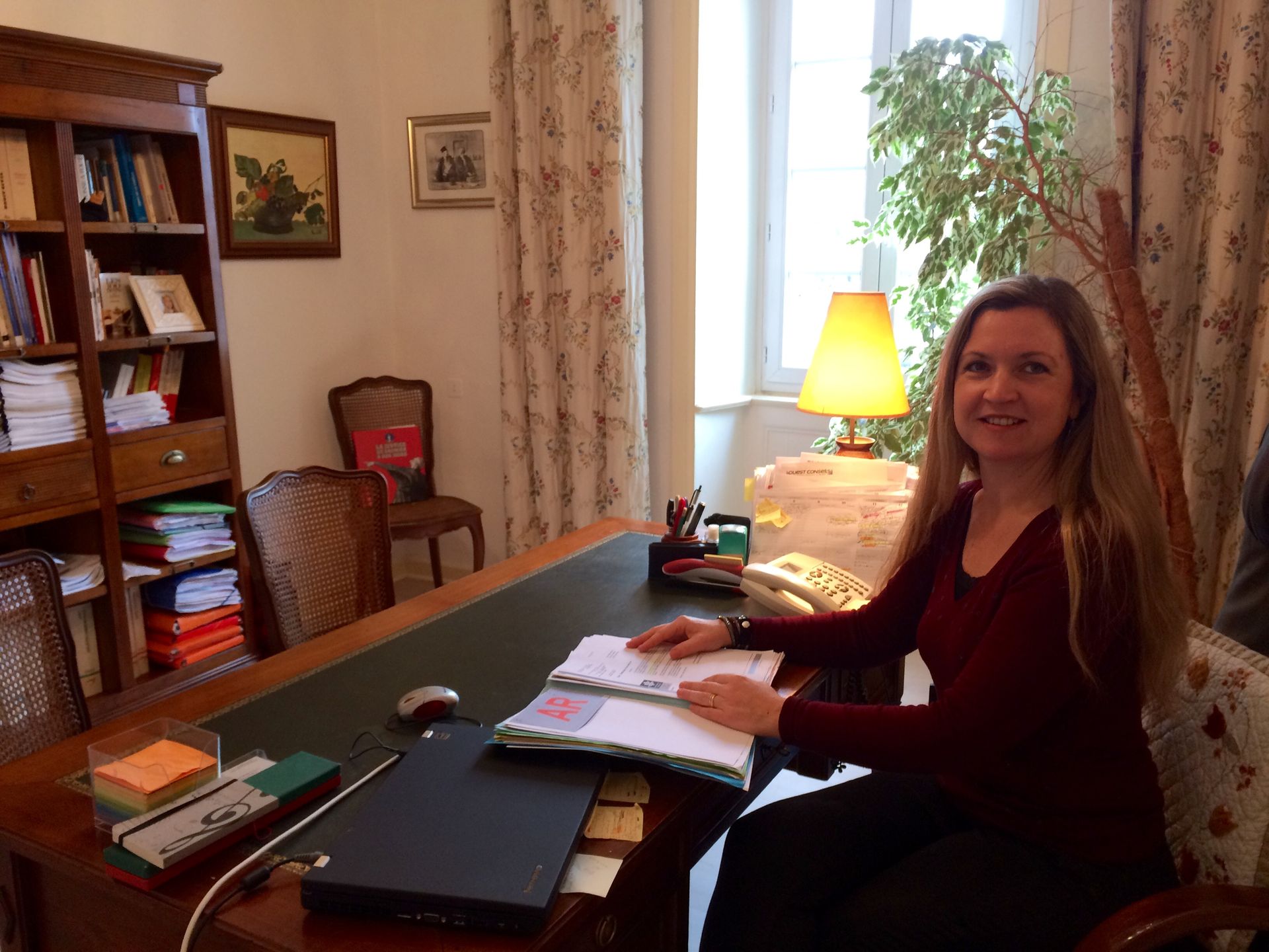 Une femme assise à un bureau dans son bureau à domicile, souriante à côté d'un ordinateur portable et de papiers, une lampe étant placée près de la fenêtre.