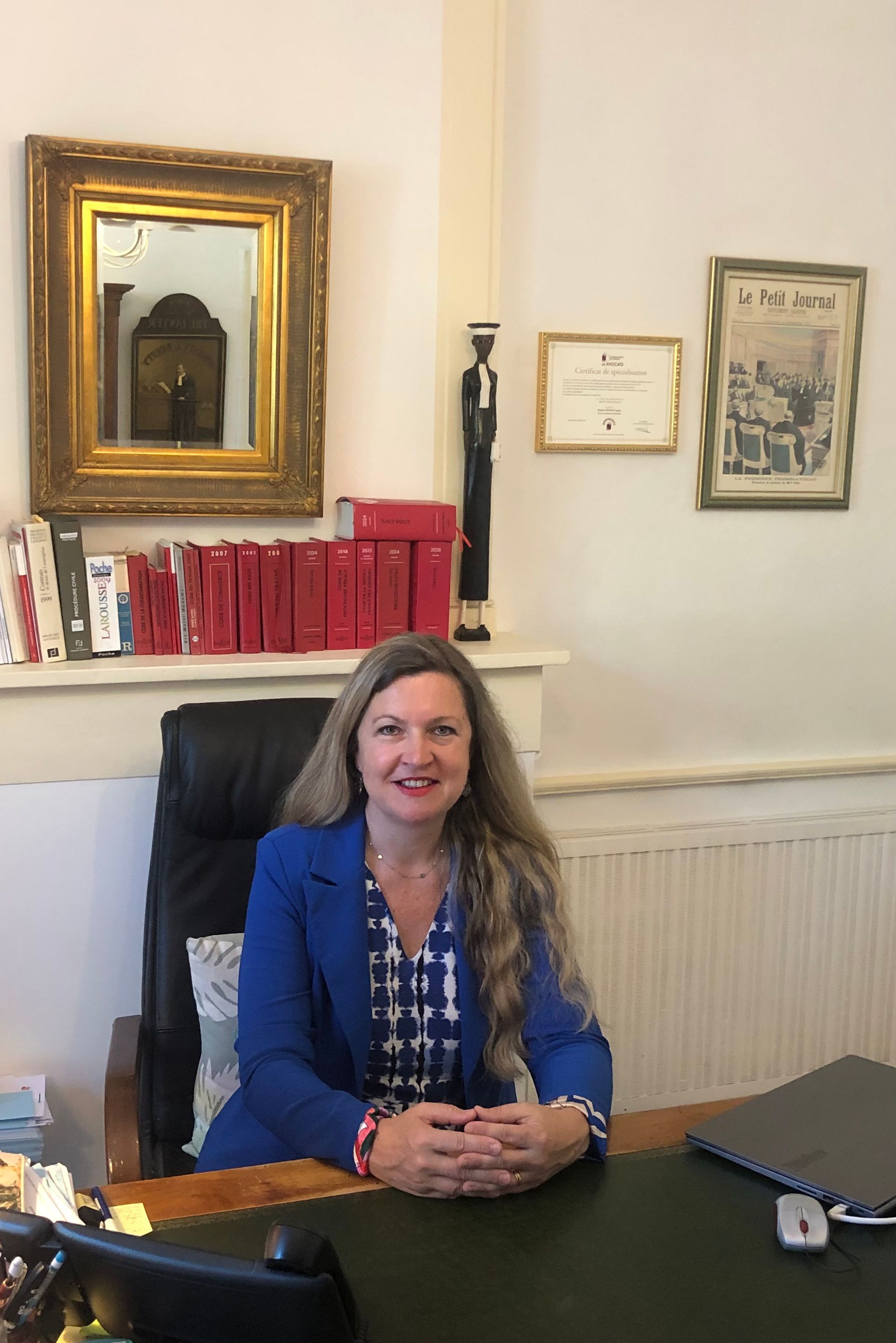 Une femme assise à un bureau, souriante, avec des tableaux encadrés et des livres derrière elle.