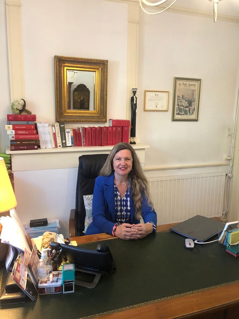 Une femme assise à un bureau, souriante, entourée de livres, d'œuvres d'art encadrées et d'une lampe.