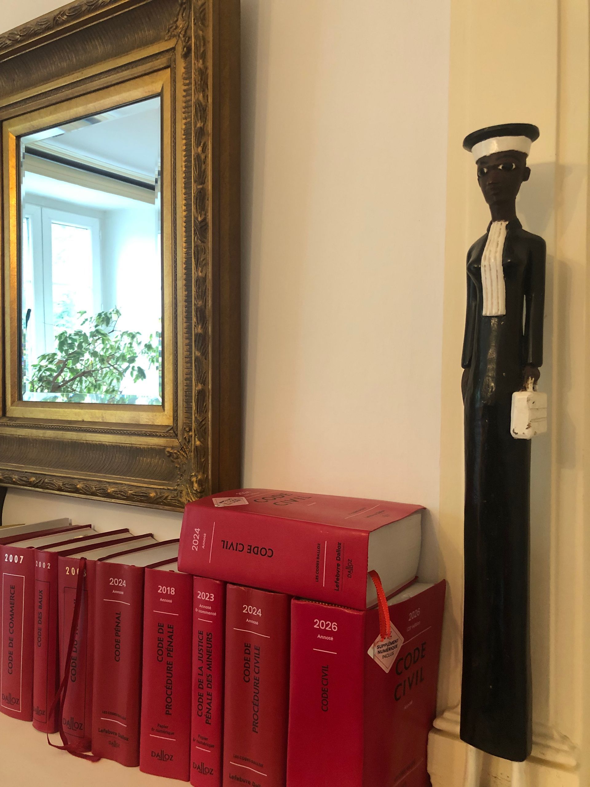 Des livres rouges alignés sur une étagère à côté d'un miroir à cadre doré et d'une grande figurine noire dans un coin.
