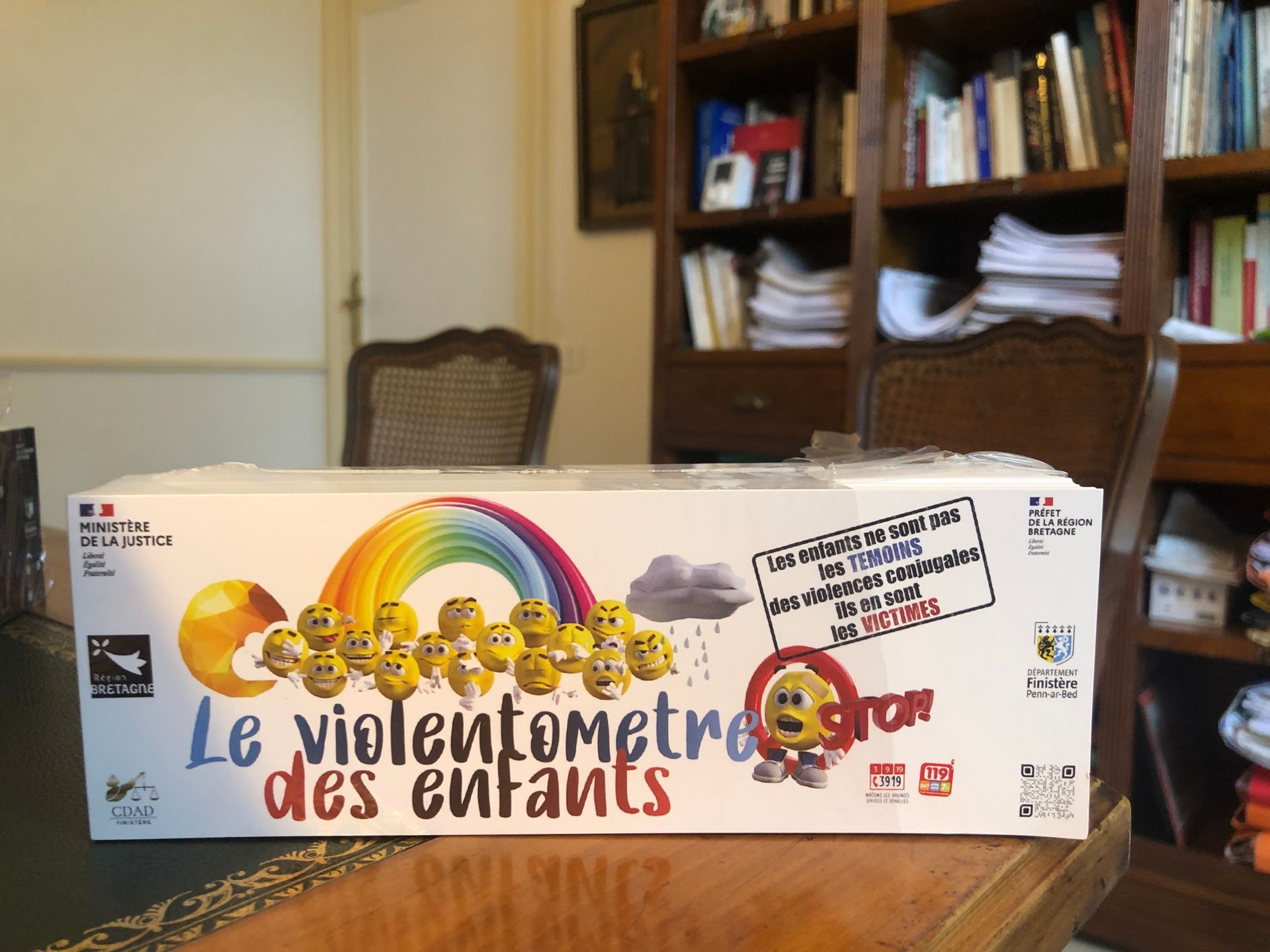 La boîte du jeu de société Le violentomètre des enfants est posée sur une table dans une pièce, avec un motif arc-en-ciel et des étagères à livres en arrière-plan.