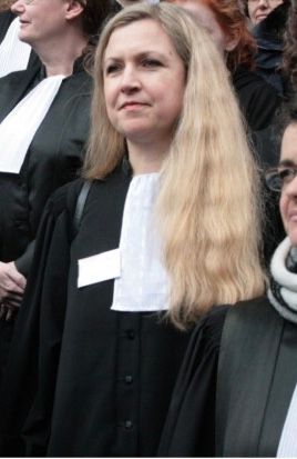 Une personne vêtue d'une robe noire à col blanc, se tenant parmi d'autres personnes à l'extérieur.