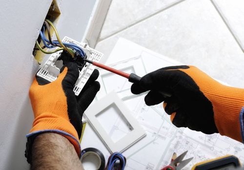 Una persona que lleva guantes naranjas está trabajando en una toma de corriente eléctrica con un destornillador.