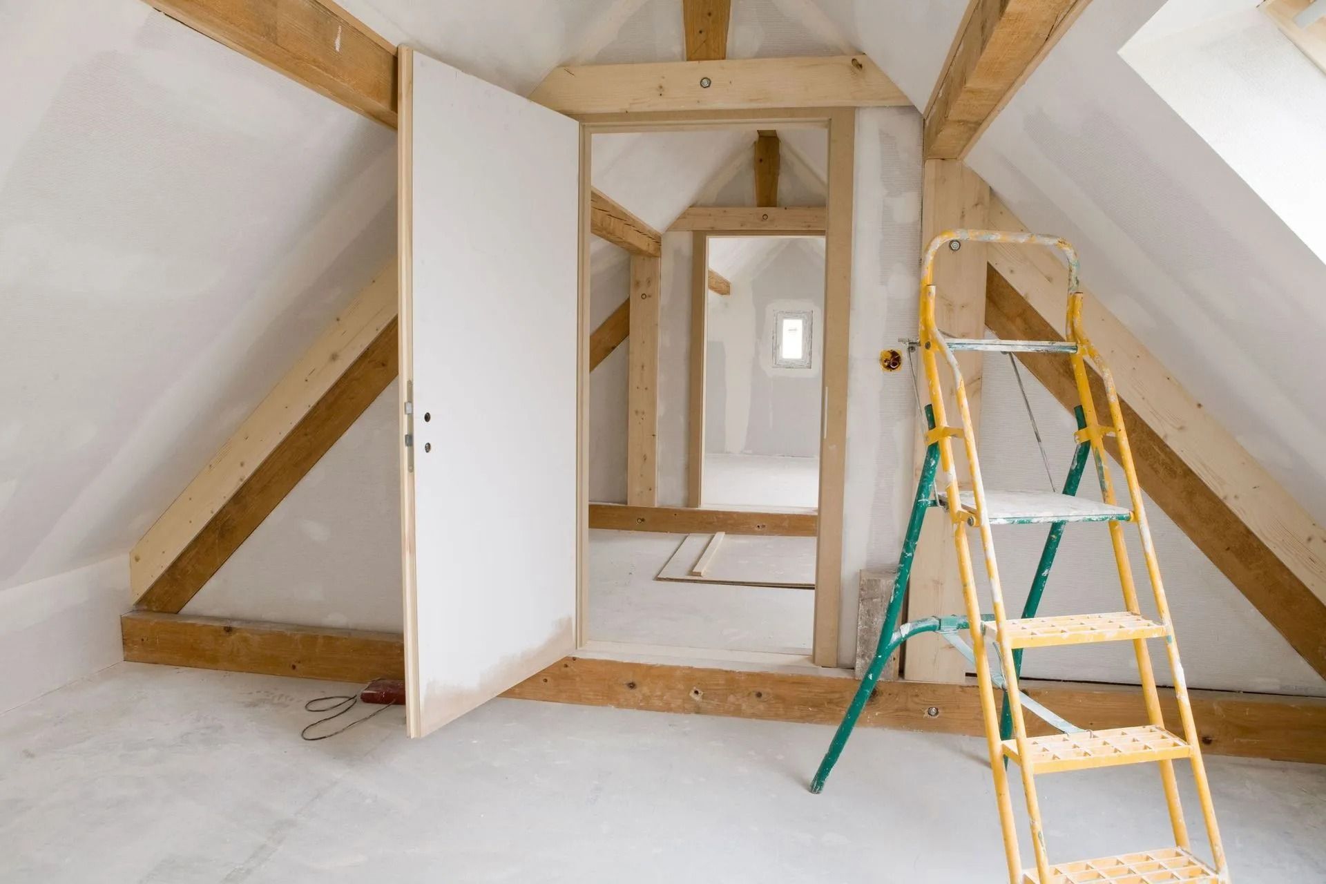 Un grenier inachevé avec des poutres en bois, des murs blancs, une porte ouverte et un escabeau jaune et vert.