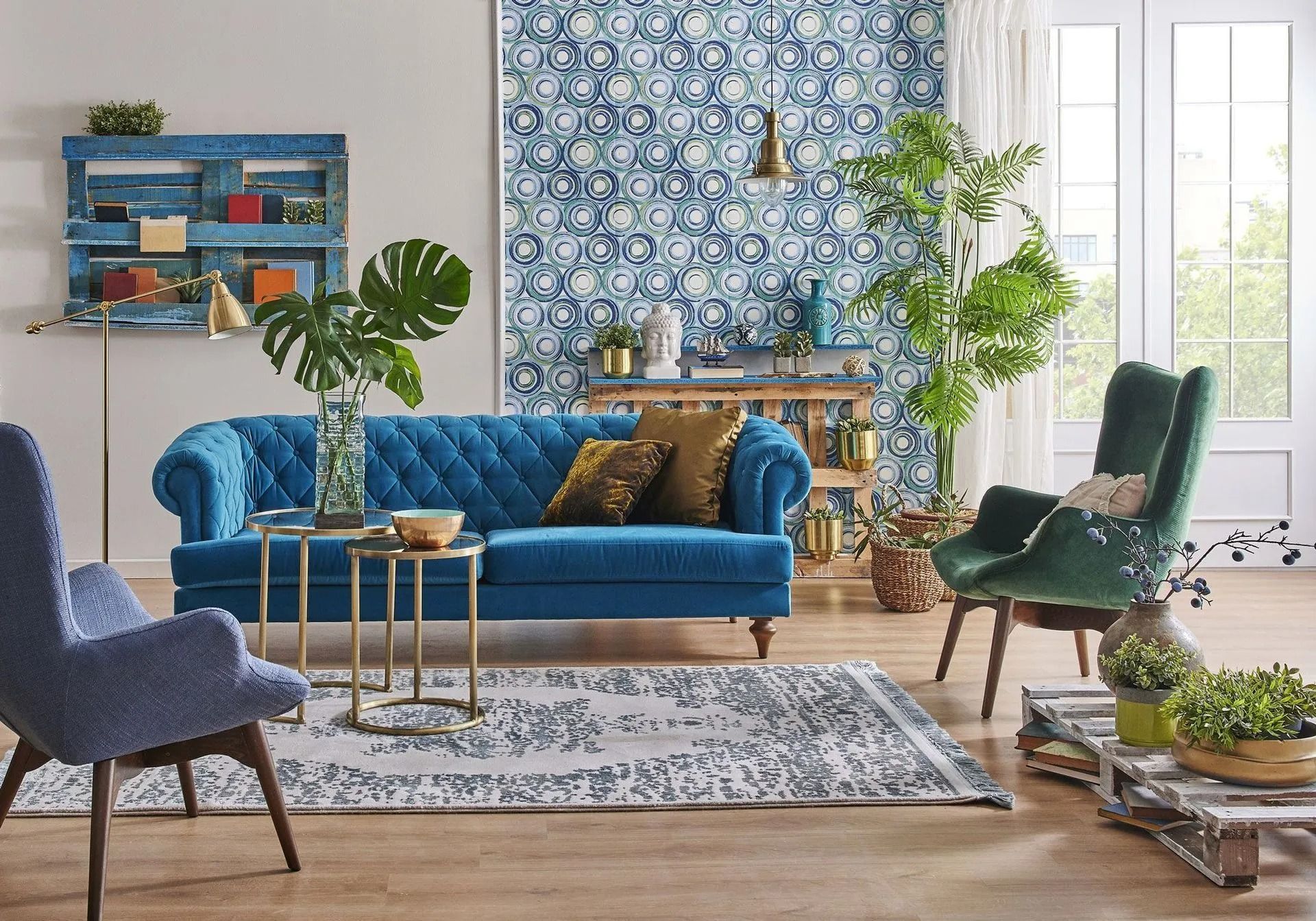 Un canapé bleu capitonné se trouve dans un salon aux murs tapissés de papier peint à motifs, agrémenté d'un fauteuil vert, de plantes d'intérieur et d'un parquet.