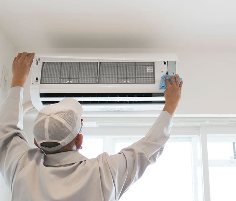 Un technicien nettoie un climatiseur mural au-dessus d'une fenêtre dans une pièce lumineuse.