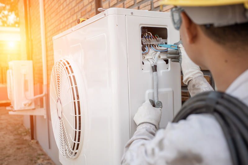 Un technicien inspecte une unité de climatisation extérieure avec ses outils, le câblage étant visible, en plein soleil.