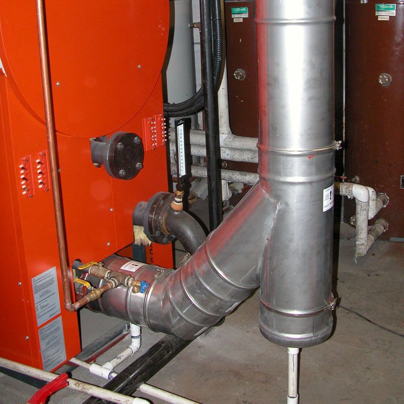 Conduits et tuyauteries industriels à l'intérieur d'un local technique, avec des équipements rouges et un grand tuyau de ventilation argenté.