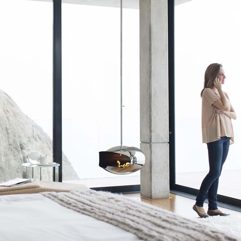 Femme debout près de grandes fenêtres dans une pièce moderne, regardant à l'extérieur près d'une cheminée suspendue.