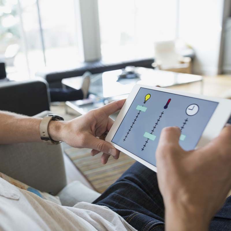 Des mains tenant une tablette affichant une interface d'application colorée sont assises sur un canapé dans un salon lumineux.