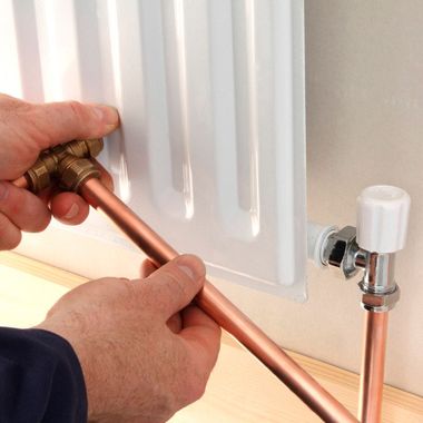 Des mains raccordent des tuyaux de cuivre à un robinet de radiateur blanc sous une fenêtre