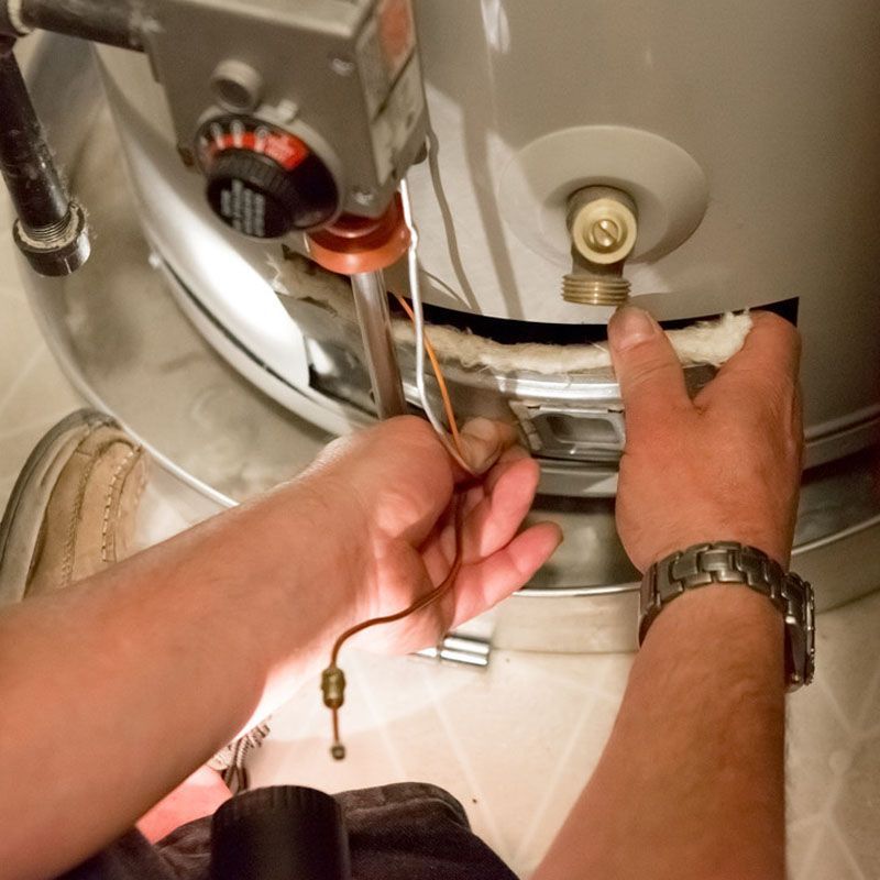 Des mains manipulent la vanne de gaz et la tuyauterie d'un chauffe-eau dans un placard technique.