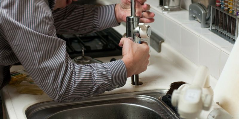 Une personne fait la vaisselle à un évier de cuisine avec eau courante et produits de nettoyage à proximité.