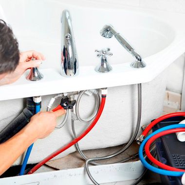 Un technicien répare la plomberie sous un lavabo blanc, avec des tuyaux rouges et bleus raccordés.