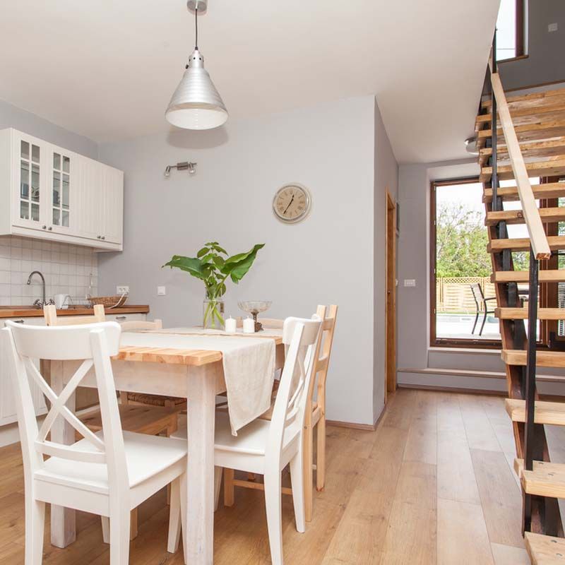 Salle à manger lumineuse avec une table en bois, des chaises blanches, des placards de cuisine, un escalier et une suspension lumineuse.