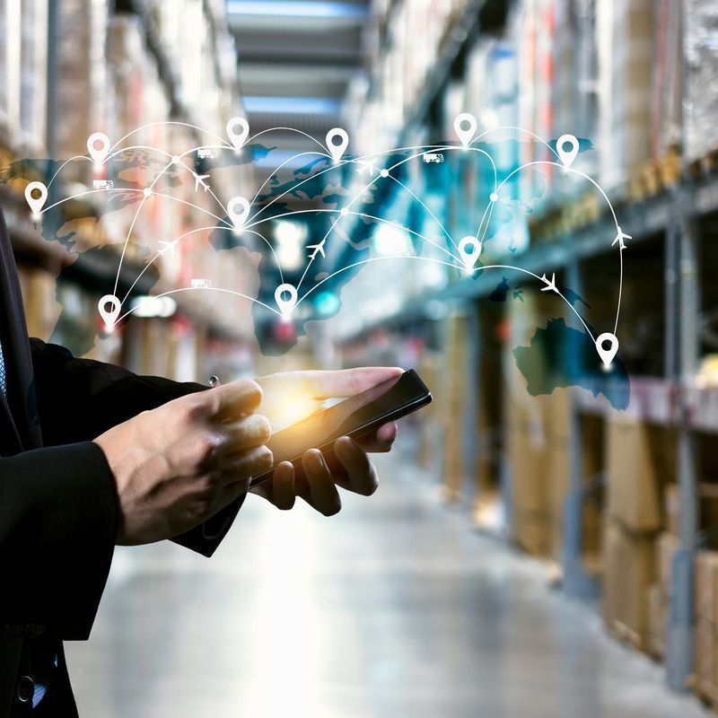 Une personne utilise une tablette dans un entrepôt, avec des icônes de réseau numérique superposées aux étagères et aux cartons.