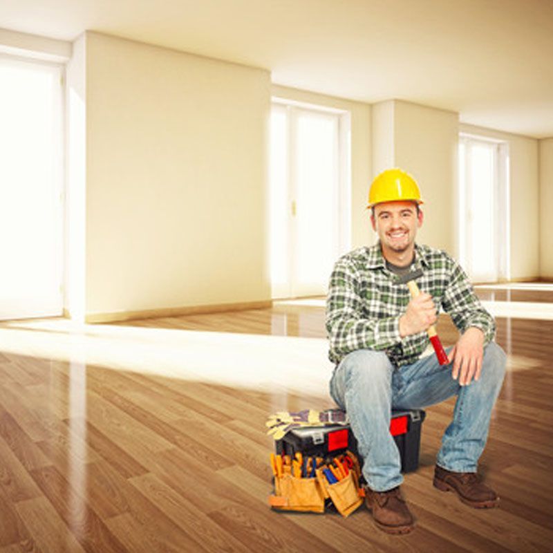 Un ouvrier portant un casque de chantier jaune est assis sur une boîte à outils dans une pièce lumineuse et vide, souriant et levant le pouce.