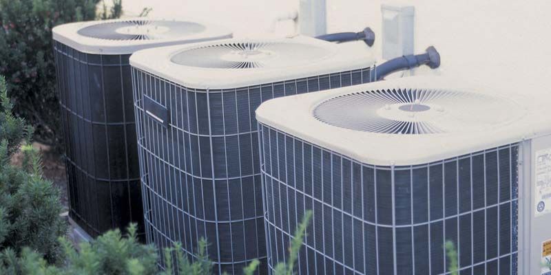 Trois grandes unités de climatisation extérieures avec ventilateurs et côtés en métal foncé, placées à côté de la végétation.