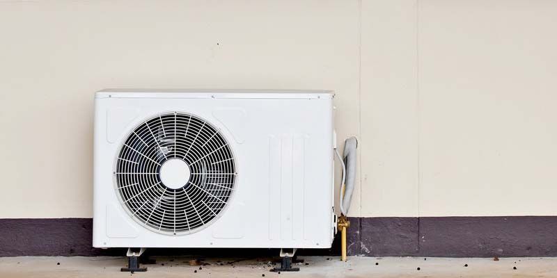 Unité de climatisation extérieure contre un mur beige avec tuyaux et câbles apparents.