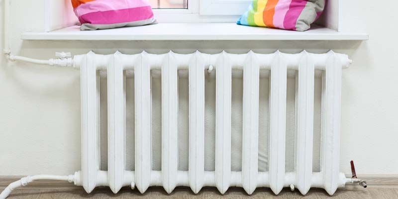 Radiateur blanc sous une fenêtre avec des coussins colorés sur le rebord.