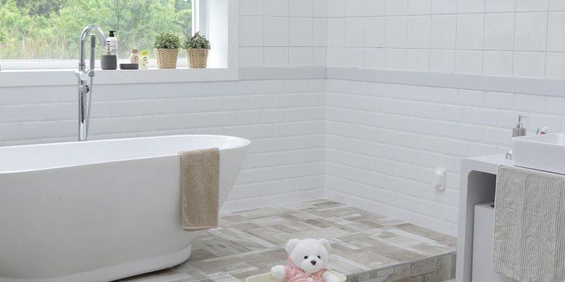 Salle de bains d'un blanc éclatant avec baignoire îlot, plantes vertes près de la fenêtre et un ours en peluche posé au sol.