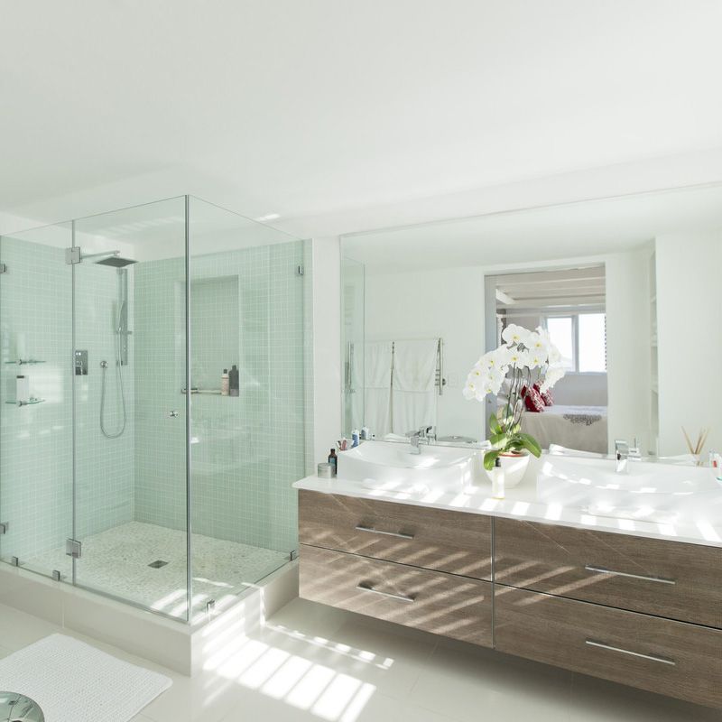 Salle de bains moderne et lumineuse avec douche en verre, double vasque et orchidée blanche près d'une fenêtre ensoleillée