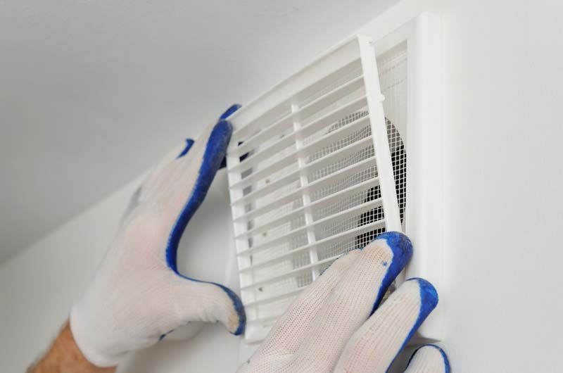 Des mains gantées installent une grille d'aération blanche sur un mur