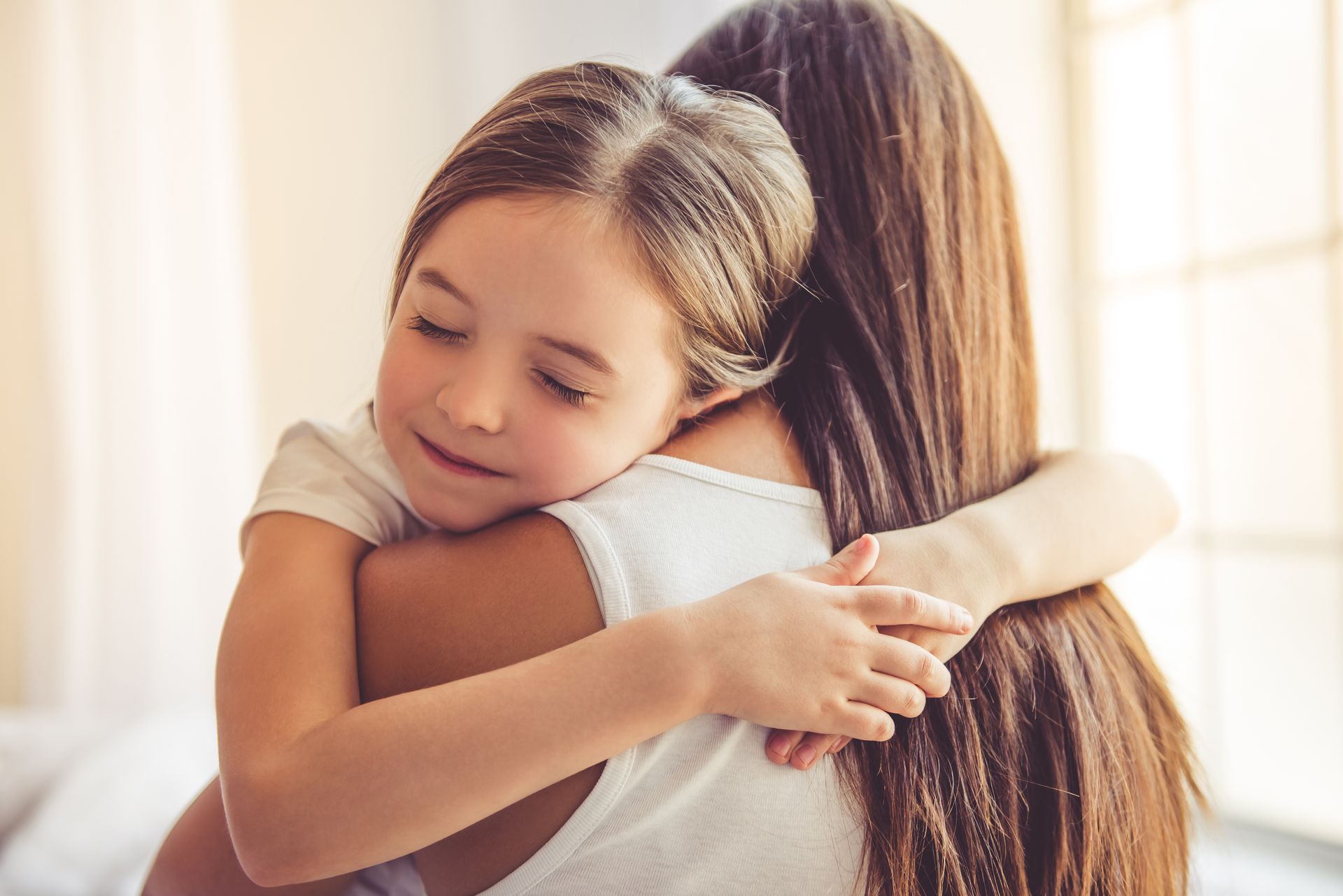 Une femme et une petite fille s'enlaçant chaleureusement près d'une fenêtre ensoleillée.