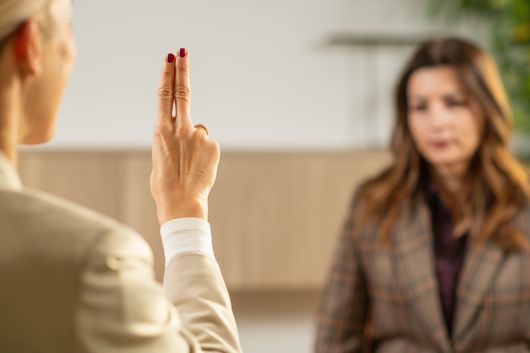 Une personne lève deux doigts lors d'une conversation avec une femme dans un décor intérieur flou.