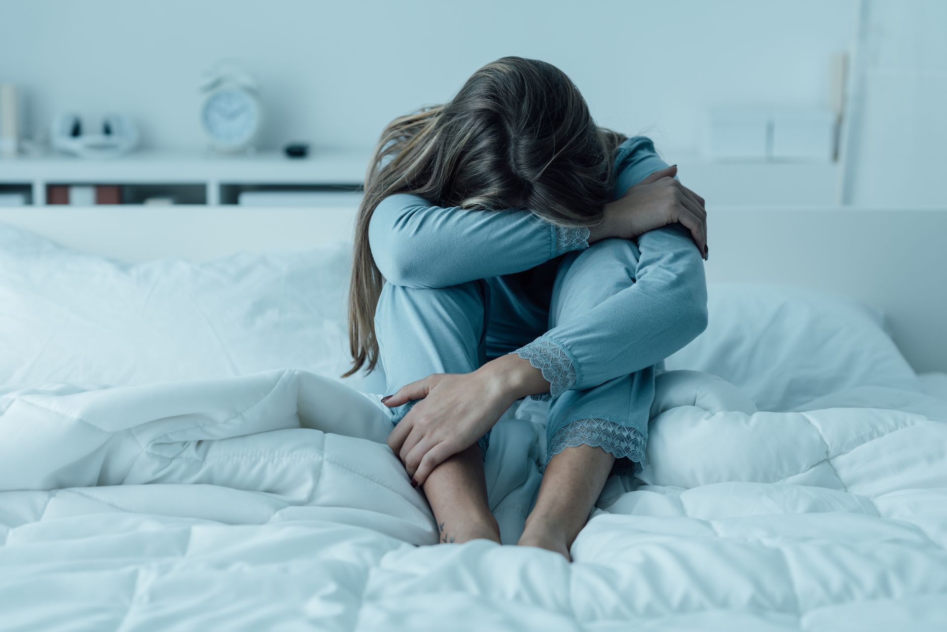 Une personne assise sur un lit, la tête baissée et les bras enroulés autour des genoux.