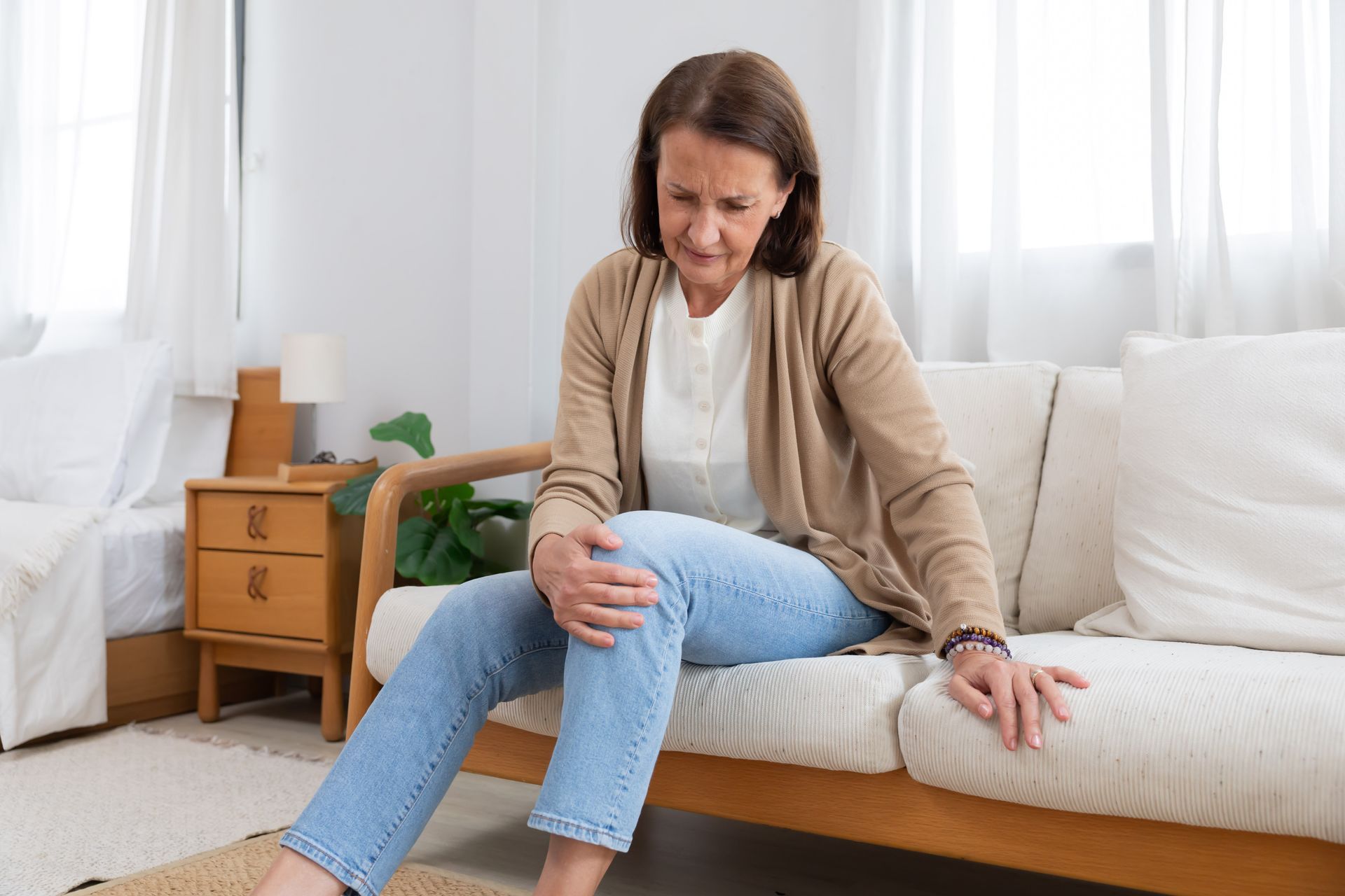 Une femme assise sur un canapé, se tenant le genou avec une expression de douleur, dans un salon lumineux.
