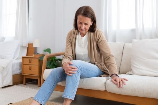 Une femme assise sur un canapé, se tenant le genou avec une expression de douleur, dans un salon lumineux.