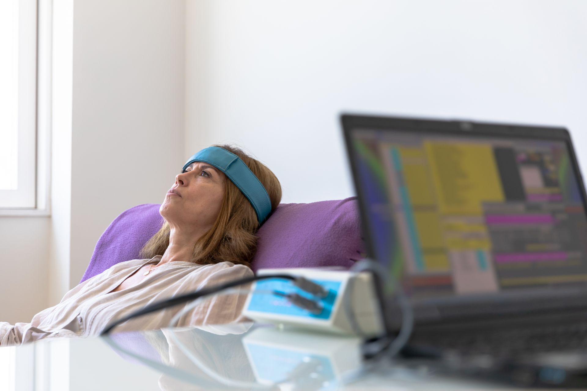 Une femme portant un casque EEG est allongée dans un fauteuil de couleur violette. Un ordinateur portable flou se trouve au premier plan.
