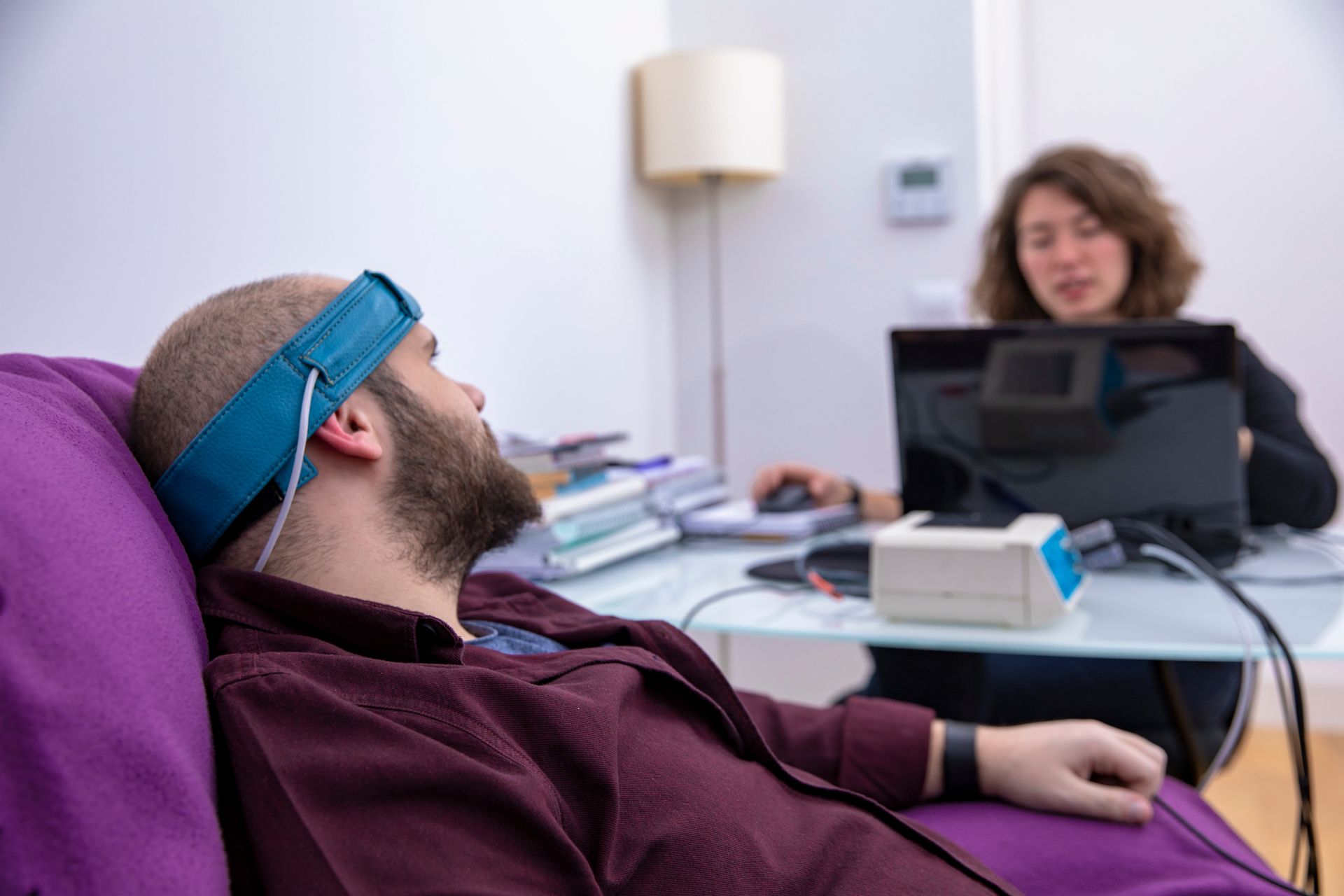 Un homme allongé sur un canapé violet avec un casque EEG sur la tête fait face à une femme assise de l'autre côté d'un bureau.