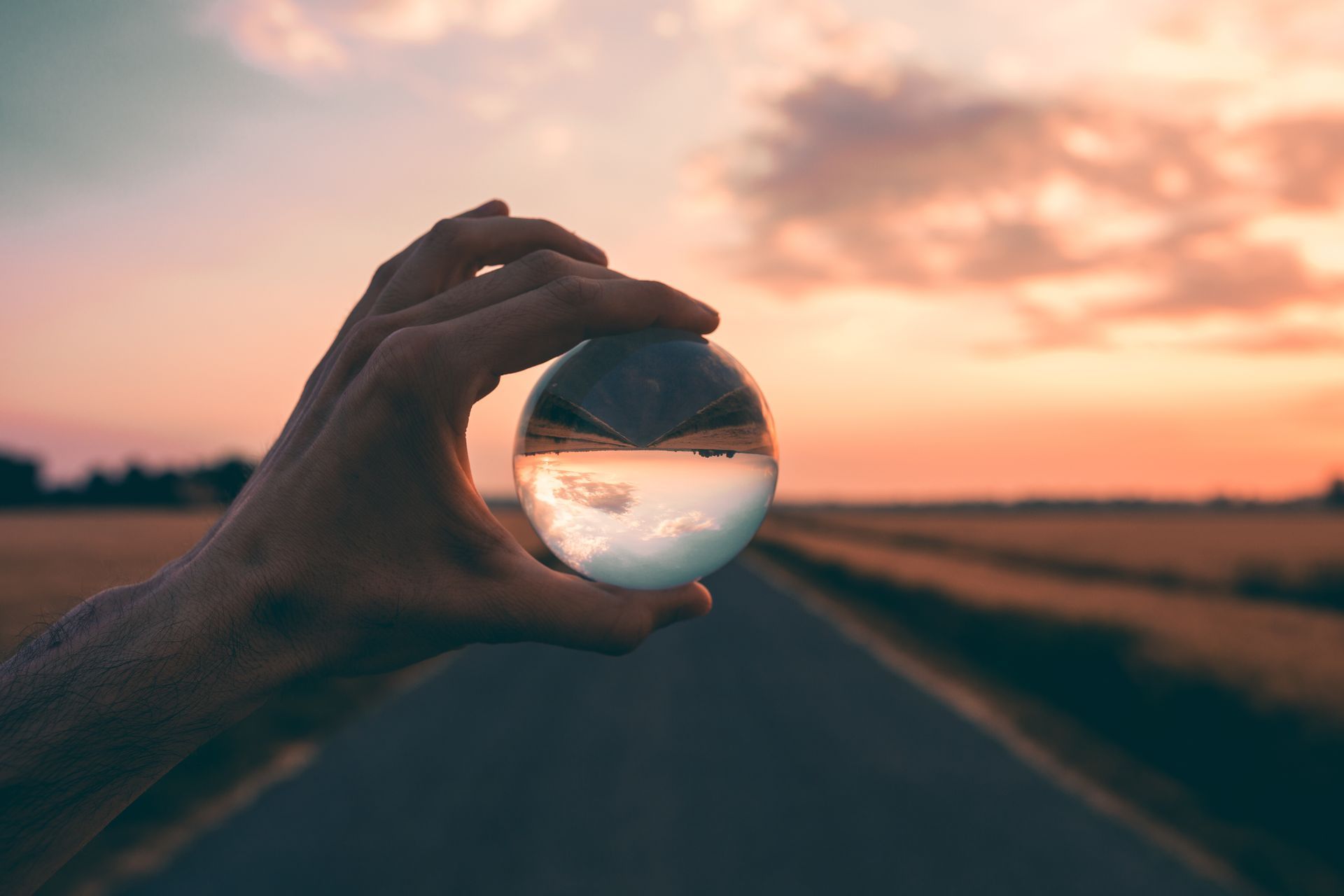 Une main tenant une sphère de verre reflétant une route au coucher du soleil, avec un horizon flou et un ciel aux couleurs chaudes.