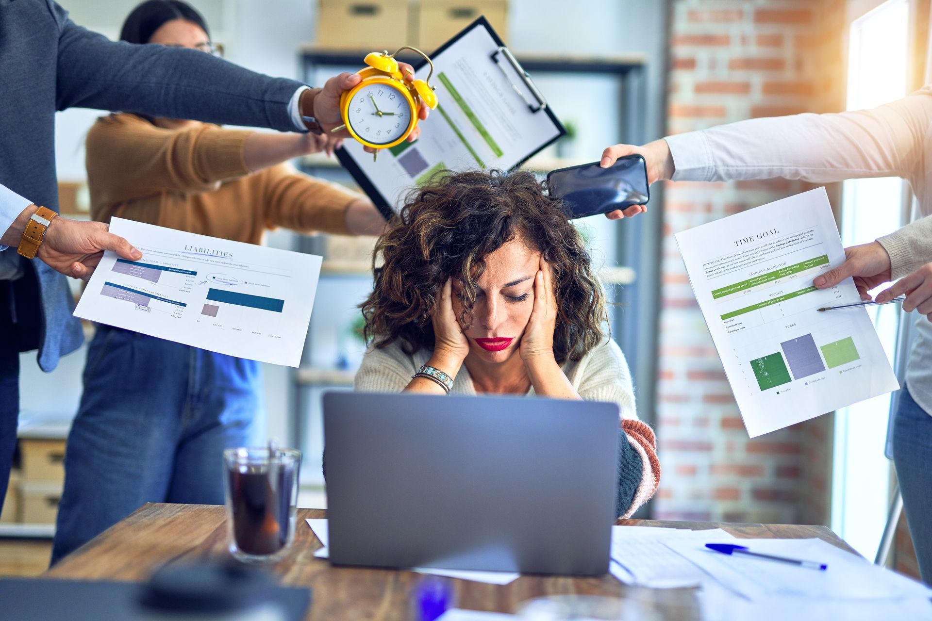 Une employée de bureau stressée devant son ordinateur portable, entourée de ses collègues chargés de papiers et de graphiques.