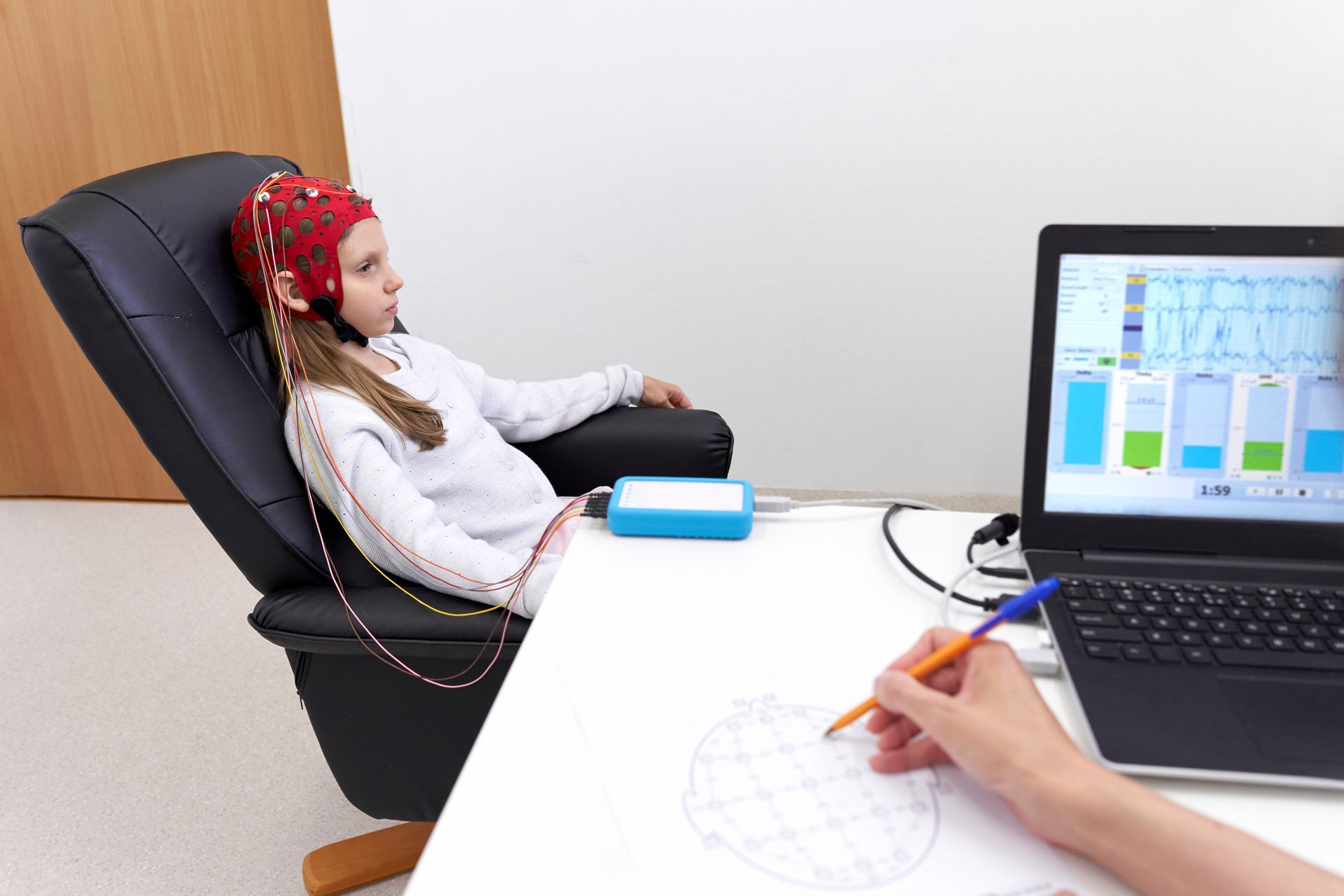 Une jeune fille portant un casque EEG est assise dans un fauteuil dans une pièce lumineuse au côté d'une professionnelle devant son ordinateur portable, prenant des notes.
