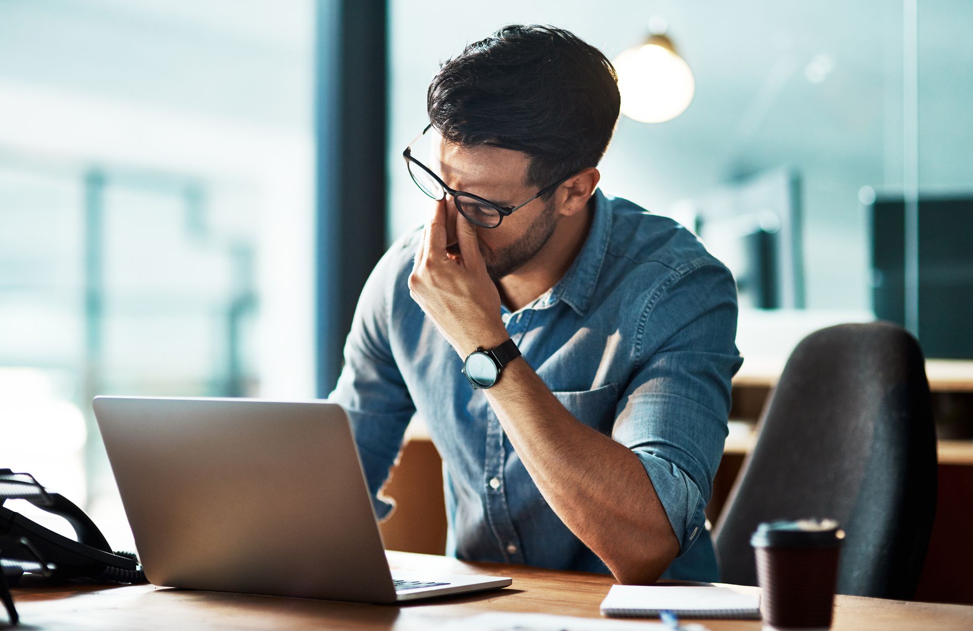 Un homme portant des lunettes se masse la tempe et regarde son ordinateur portable dans un bureau, une tasse de café posée sur le bureau.