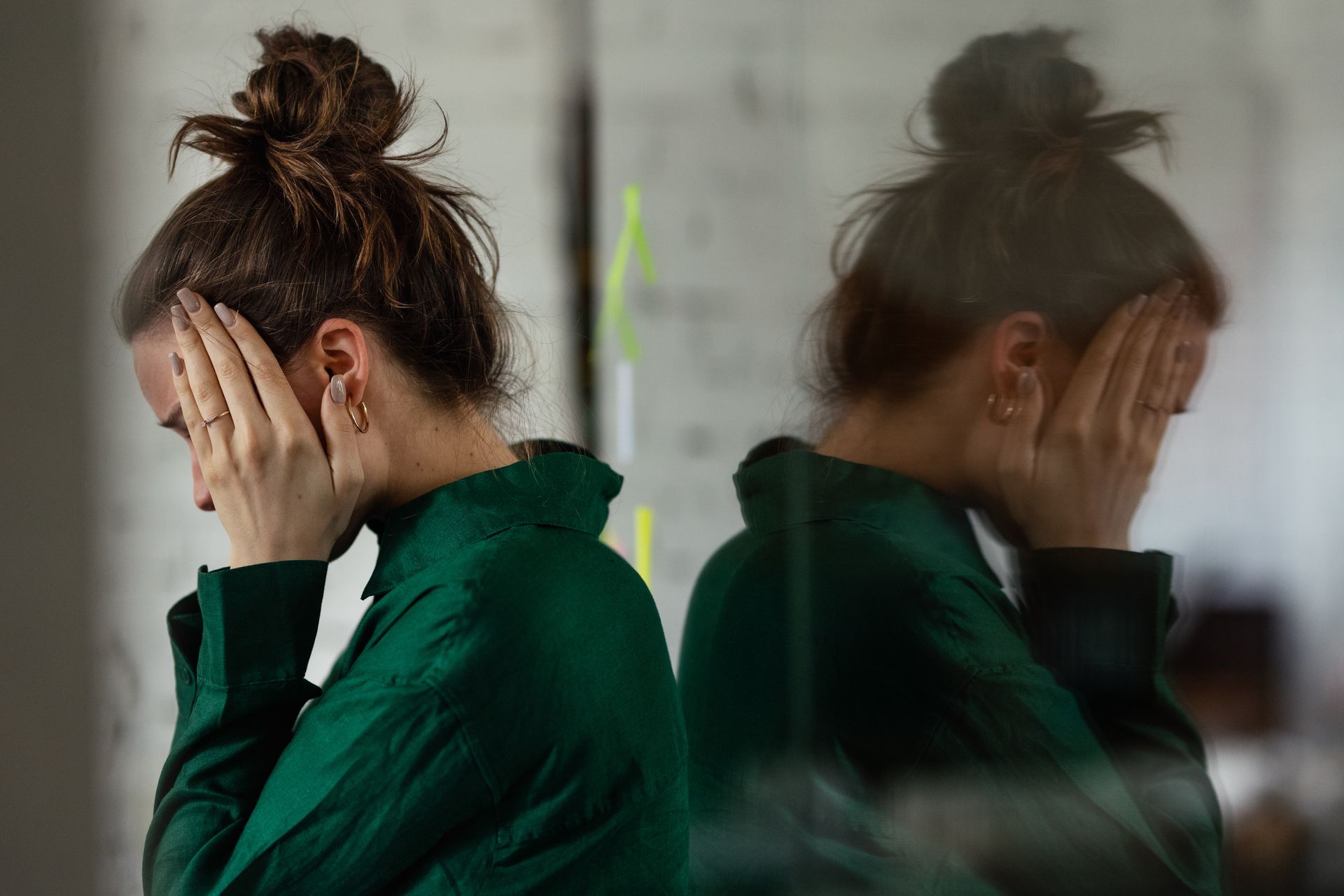 Une personne en pull vert, les mains sur son visage, est adossée à une vitre réfléchissante.