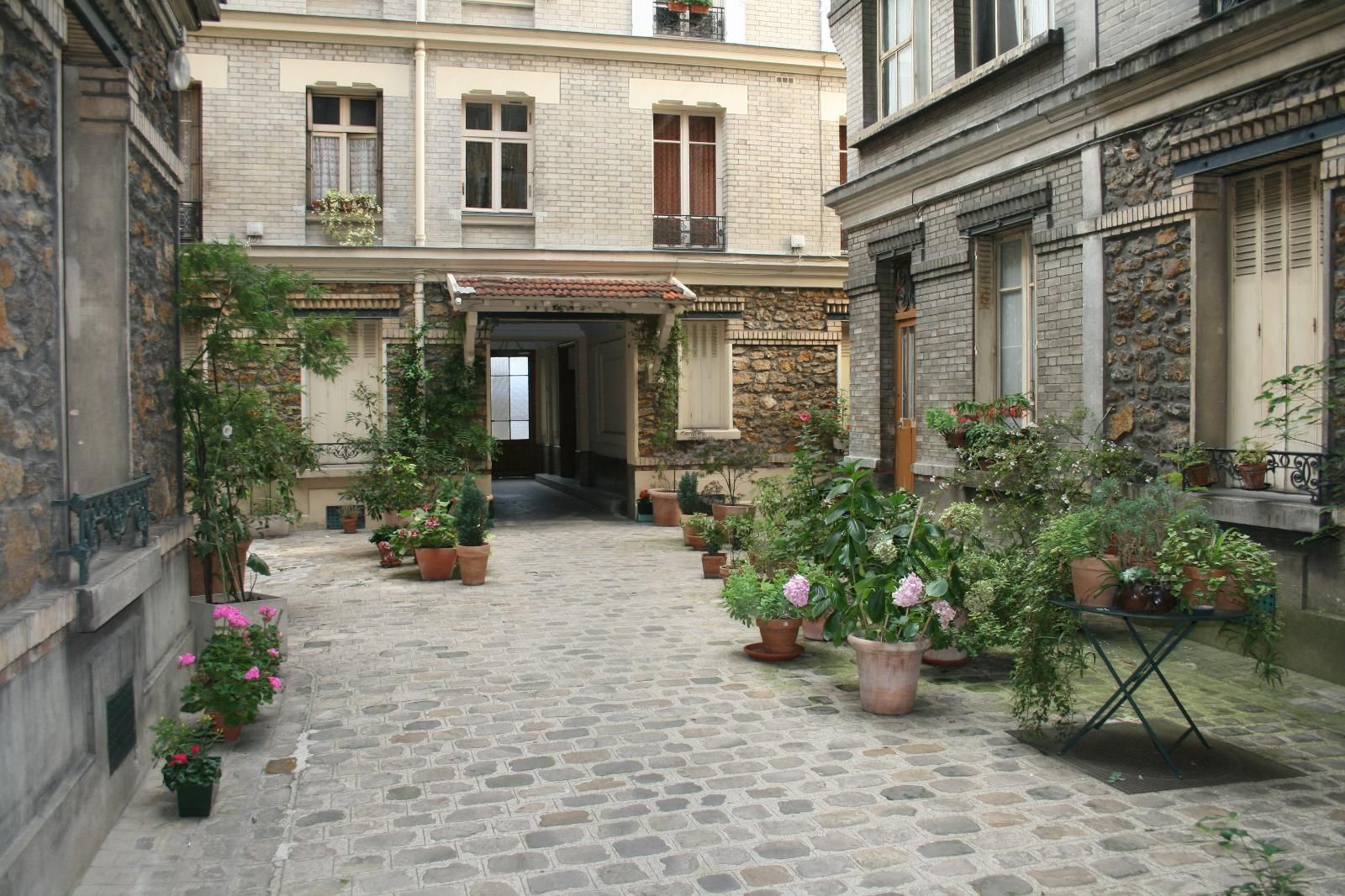 Cour pavée avec des fleurs en pot entre des bâtiments en pierre et un passage ouvert.
