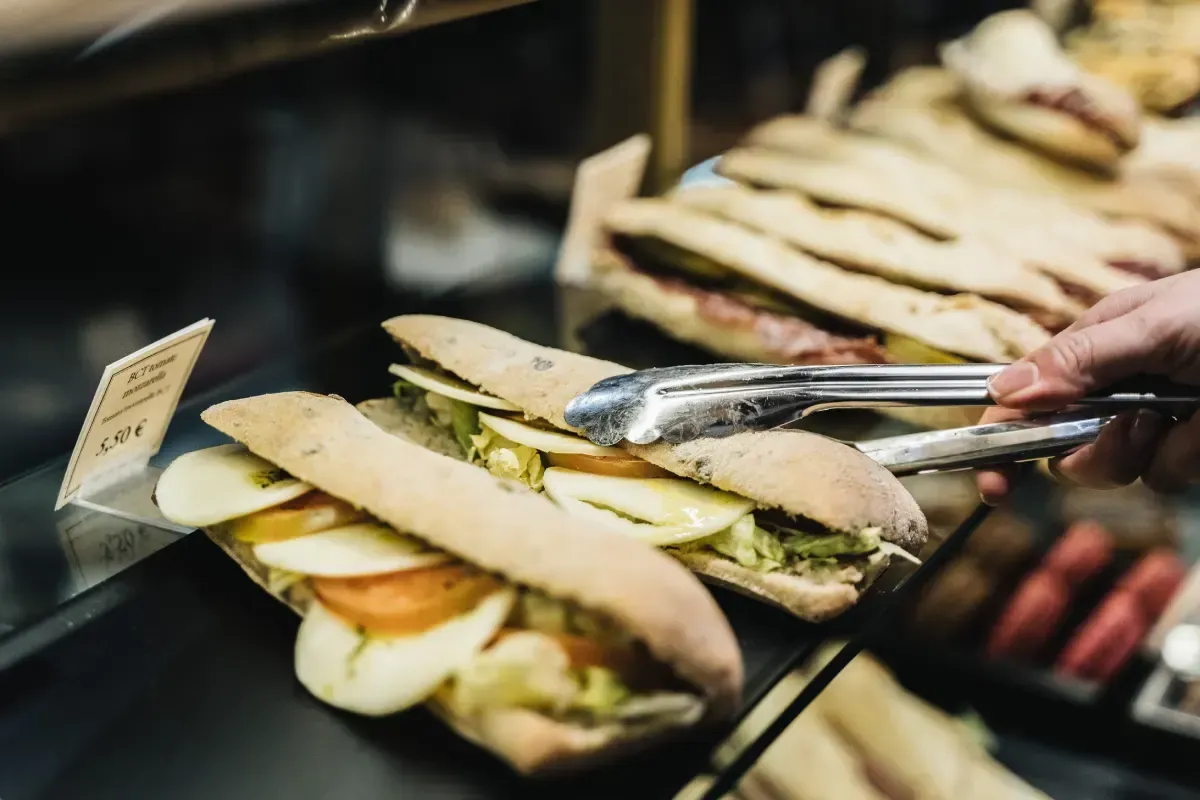 Una mano usa pinzas para sacar un sándwich de una vitrina. Se ven sándwiches con manzana y otros rellenos.