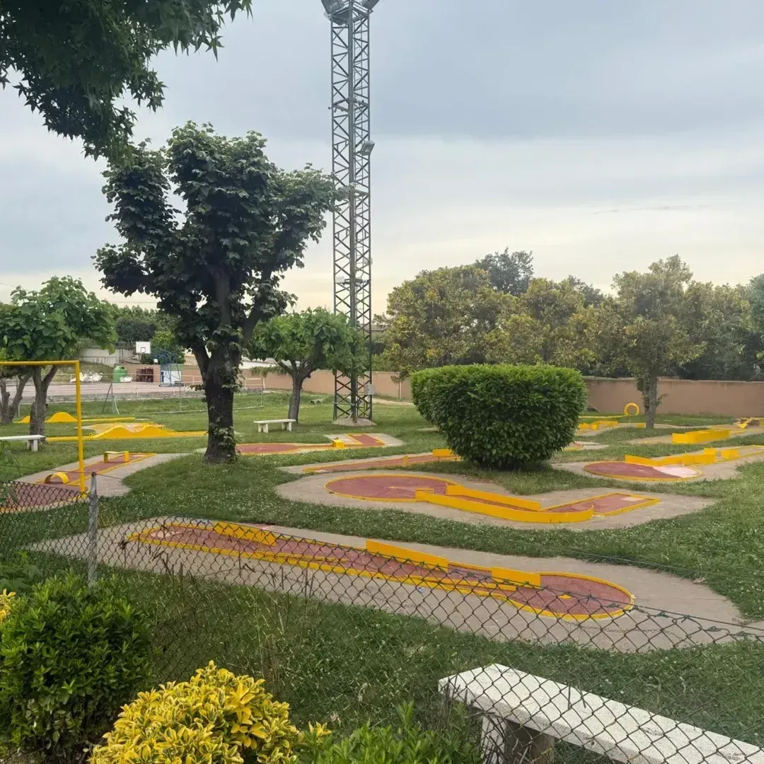 Mini campo de golf con caminos amarillos y rojos, árboles y una torre alta bajo un cielo nublado.