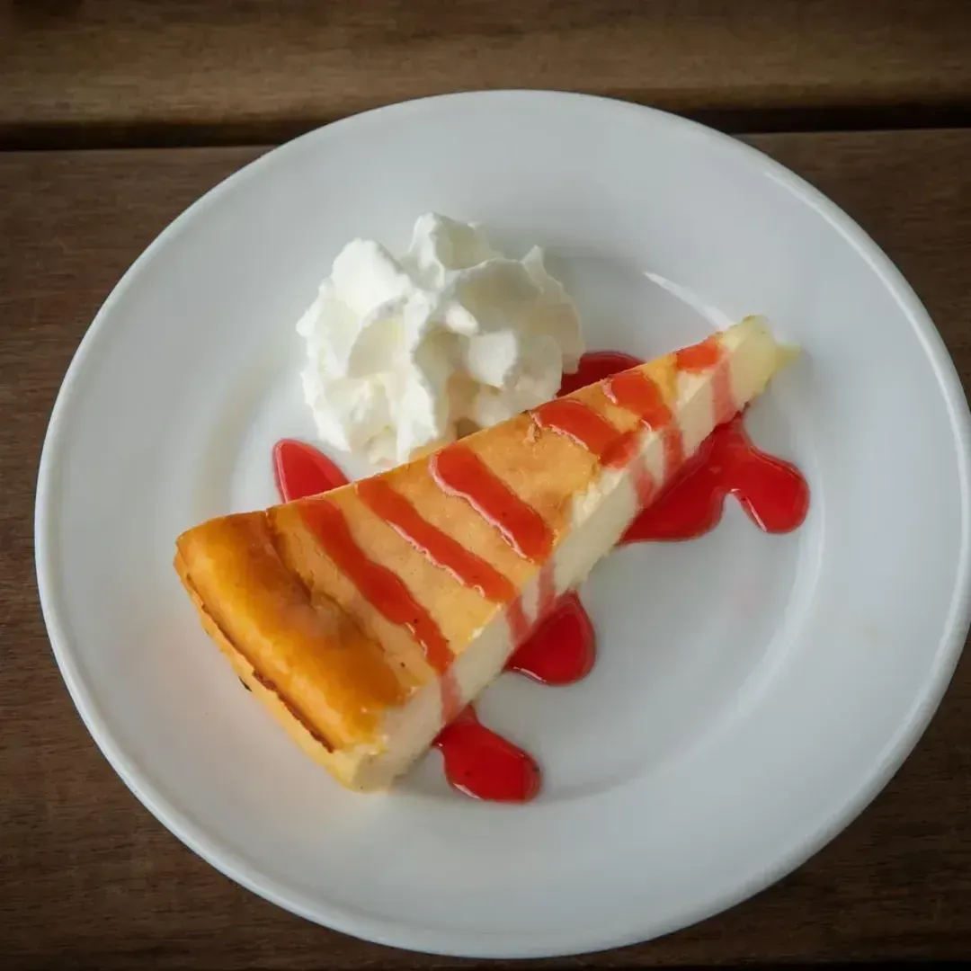 Rebanada de tarta de queso con salsa roja y crema batida en plato blanco.