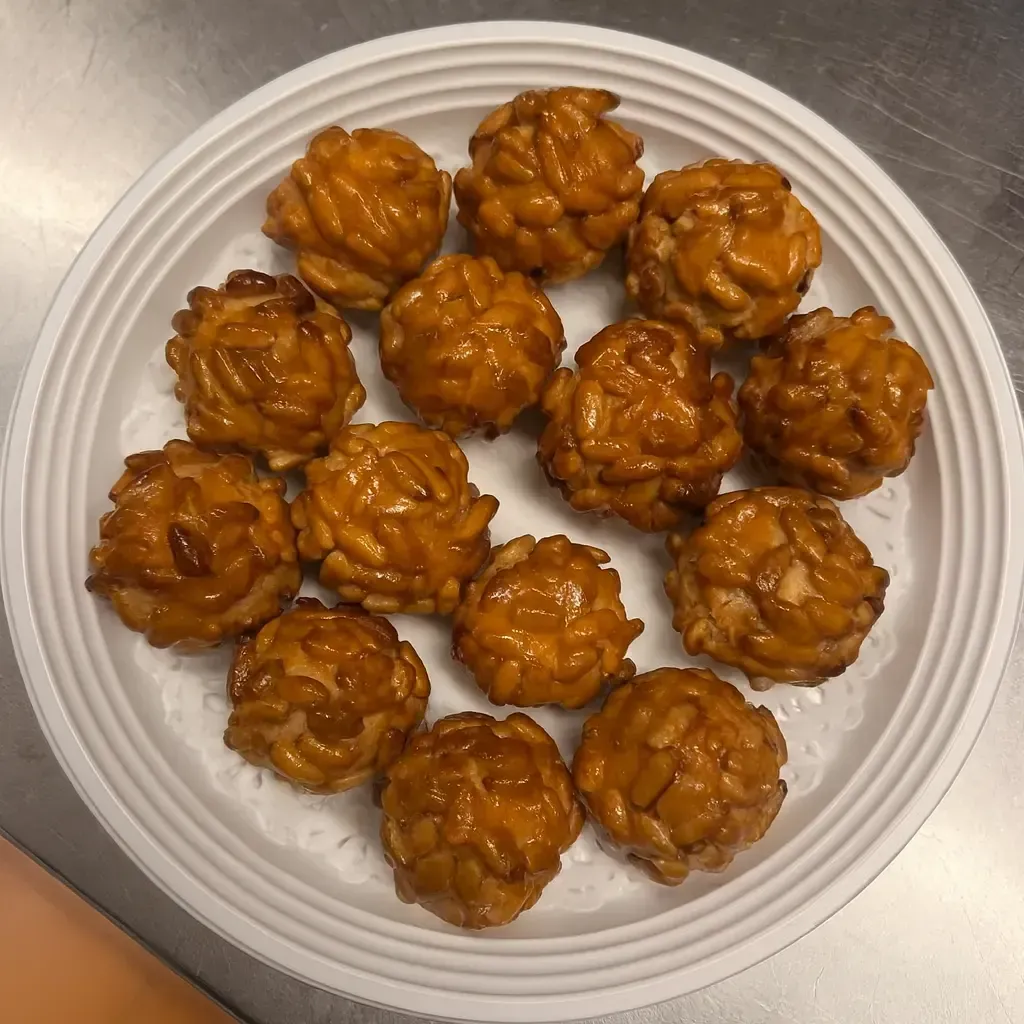 Primer plano de un plato blanco con doce panellets catalanes, de color marrón dorado, elaborados con mazapán y piñones.