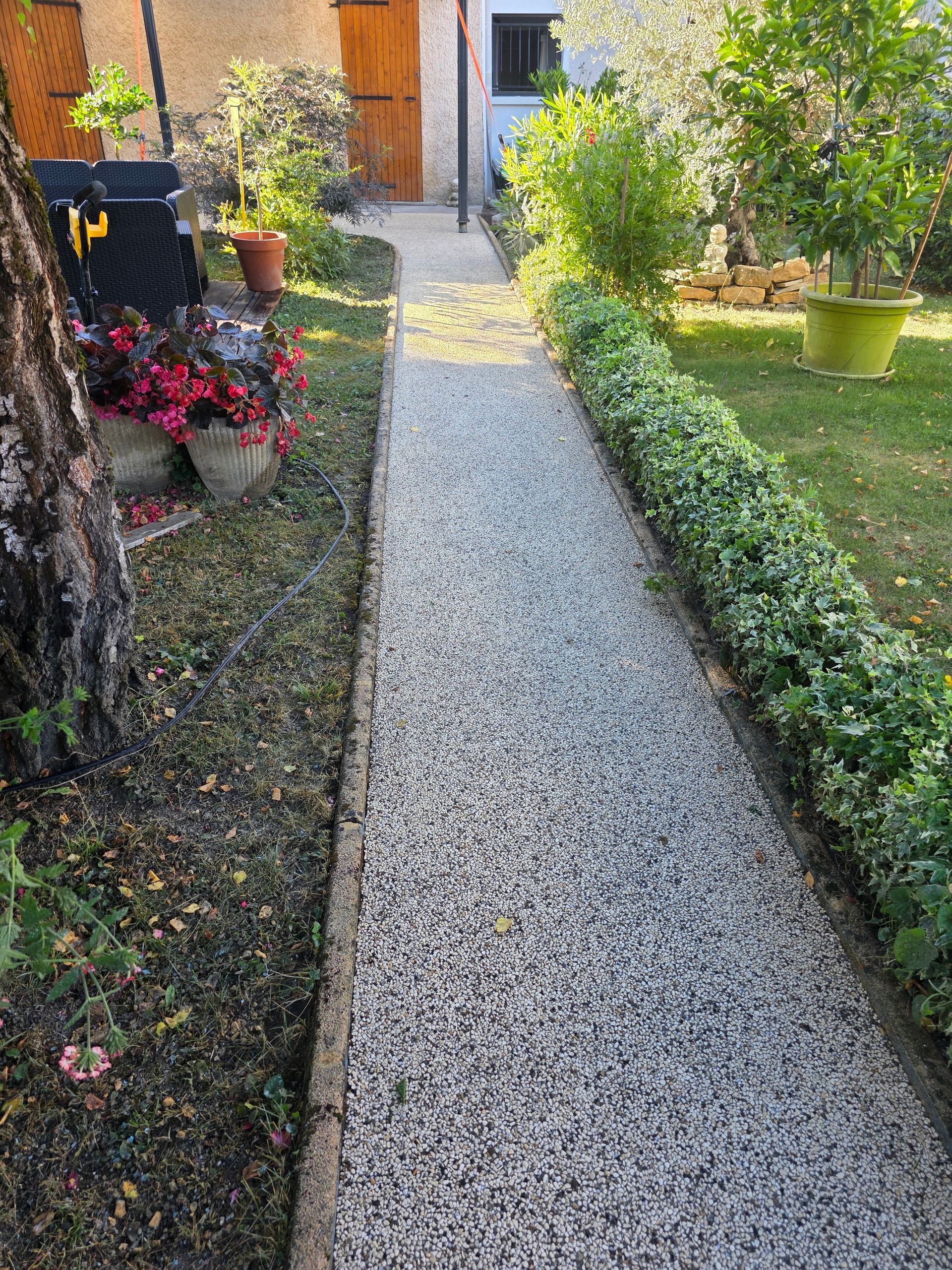 Allée de gravier menant à une porte, bordée de haies et de plantes en pot.