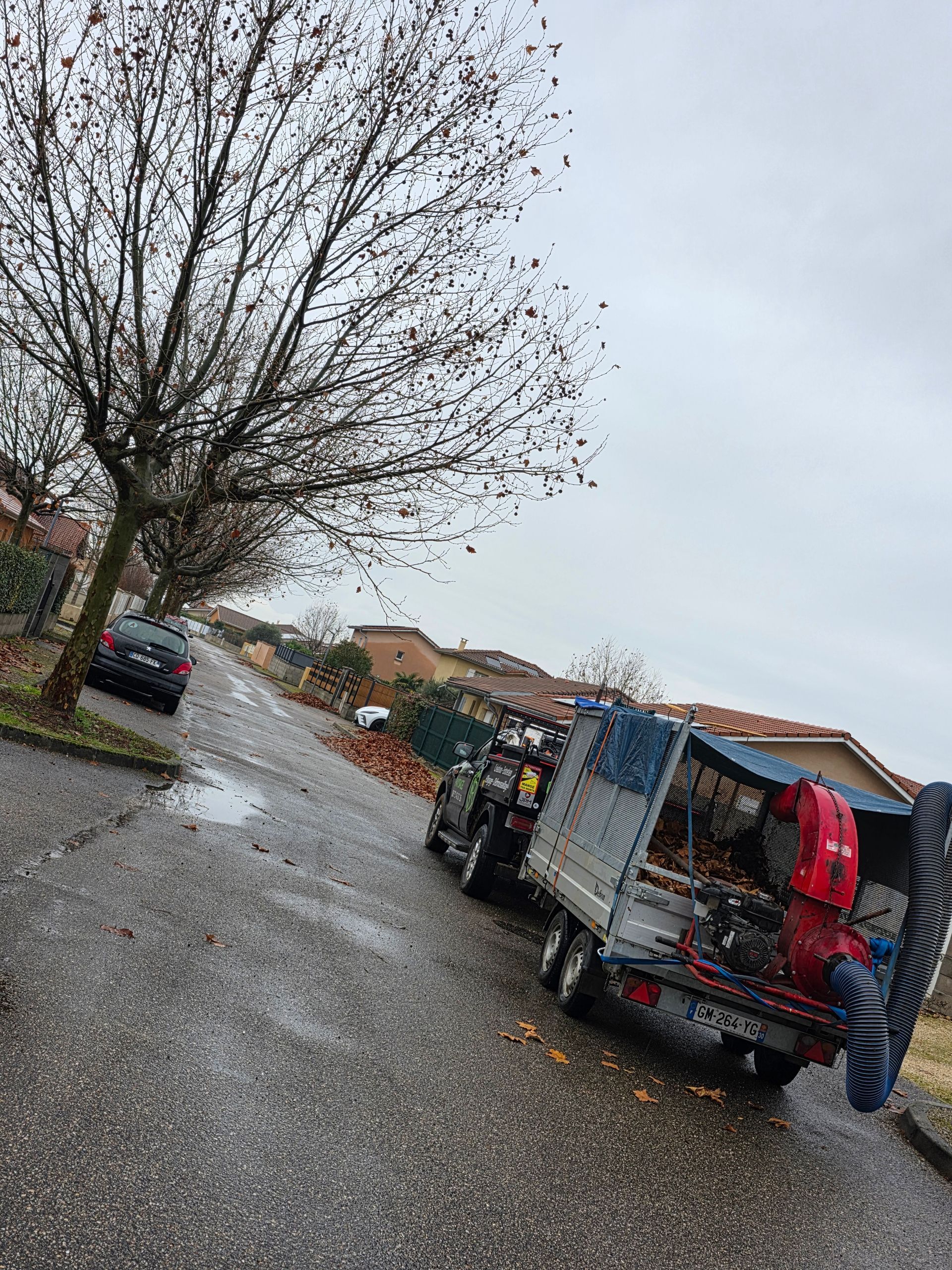 Bord de route pluvieux avec un arbre, des véhicules stationnés et une personne près d'un camion bâché dans une rue de village.