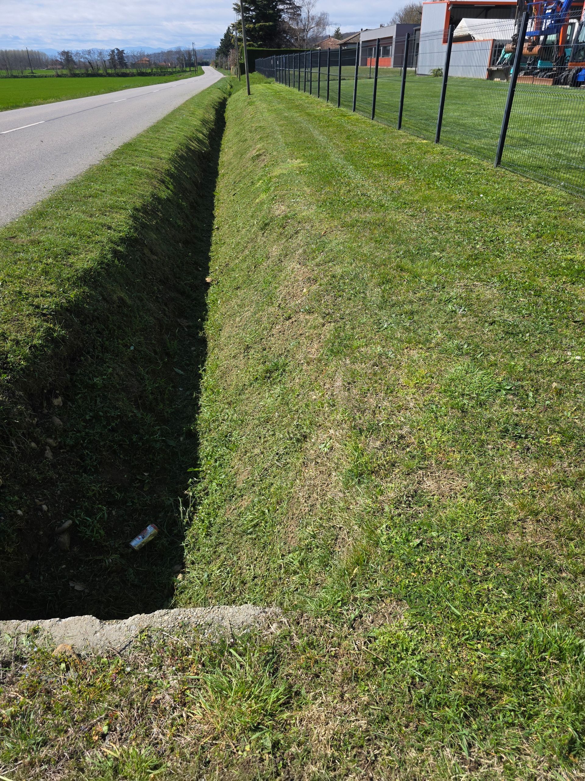 Fossé de drainage herbeux en bordure de route goudronnée et de clôture en grillage par une journée ensoleillée.